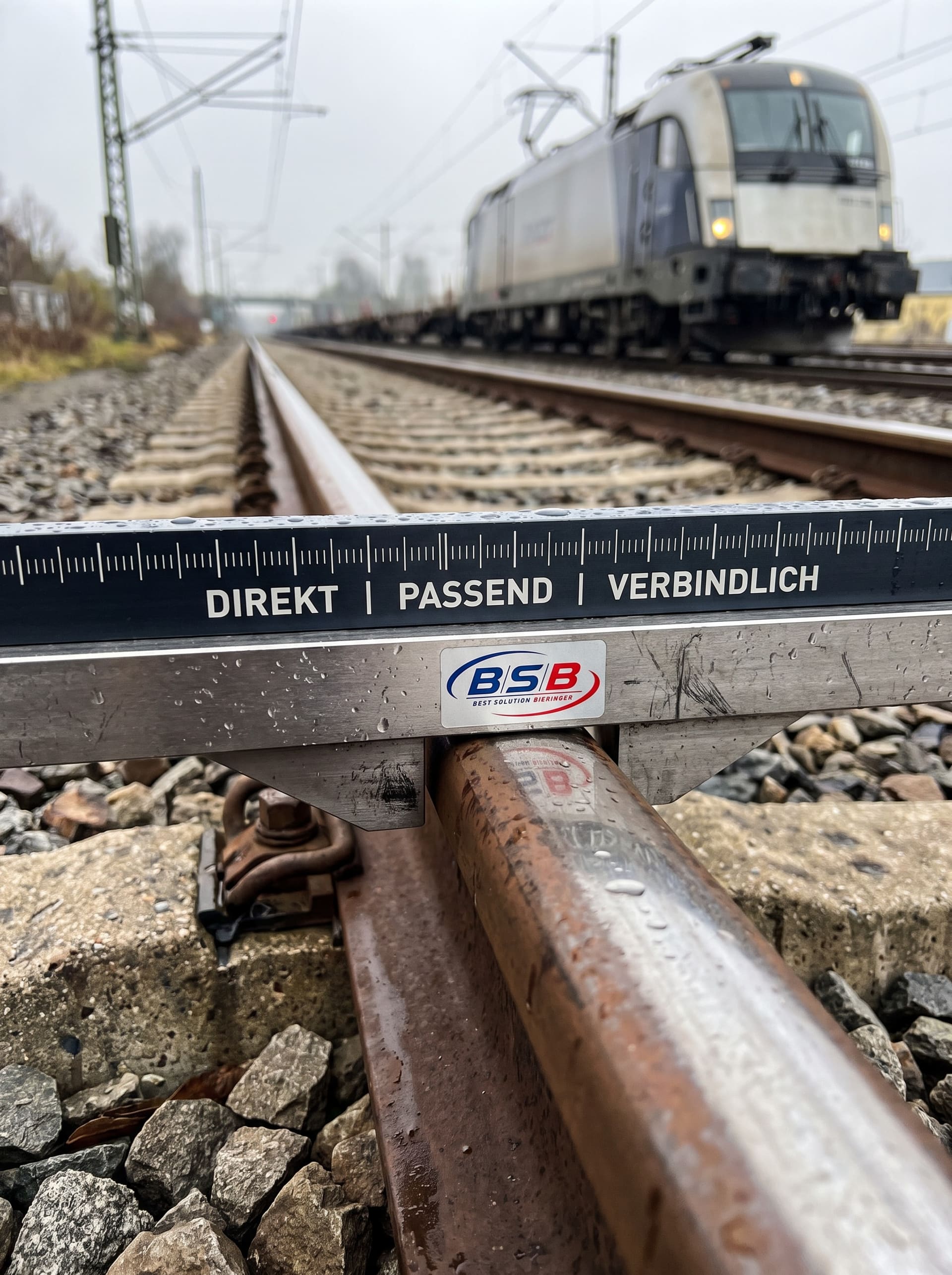 Spurweiten-Messgerät mit BSB-Logo und den Worten Direkt, Passend, Verbindlich quer über einem Gleis, dahinter eine Lokomotive
